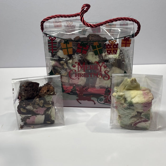 Clear Christmas gift bag with red rope handles and festive Merry Christmas decorations, containing a large 320g piece of rocky road with chocolate, marshmallows, nuts and jubes. Smaller pieces of rocky road also shown.