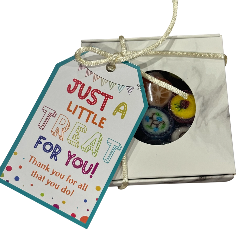 A square gift box tied with string and featuring a colorful printed tag that reads 'JUST A LITTLE TREAT FOR YOU!'. A round window on the box reveals fruit rock candy inside.
