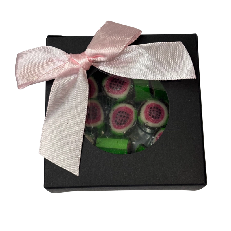 A square gift box with a round clear window showing green and pink watermelon rock candy inside, decorated with a bow.